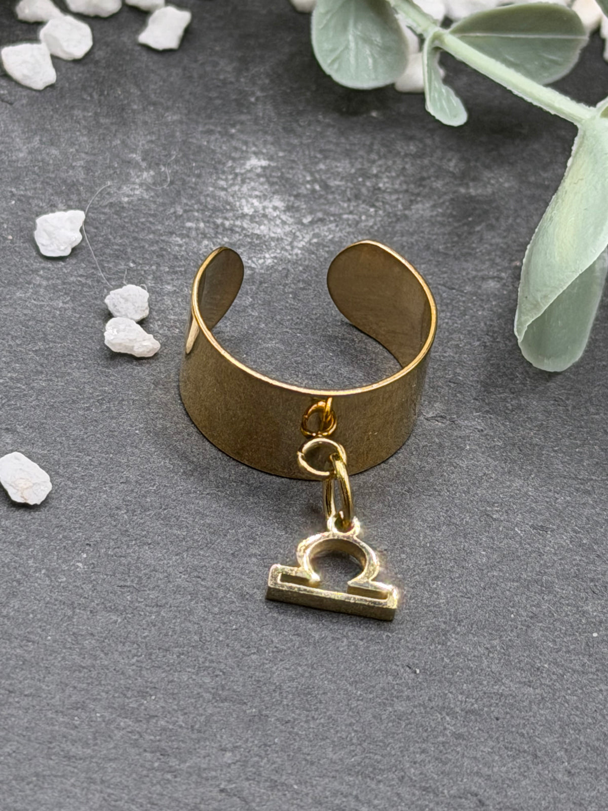 Bague Cassiopée à monture large en acier inoxydable doré, avec une breloque du signe de la Balance suspendue.