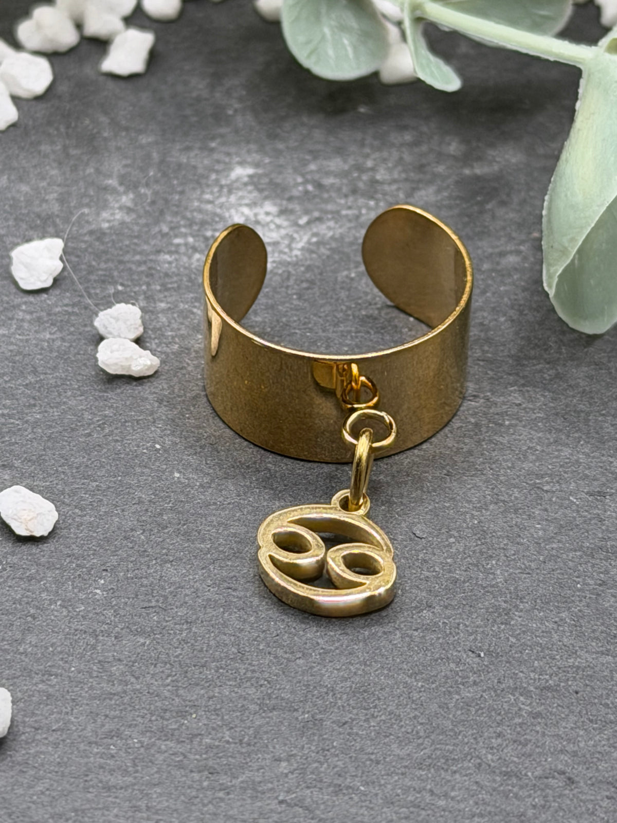 Bague Cassiopée à monture large en acier inoxydable doré, avec une breloque du signe du Cancer suspendue.