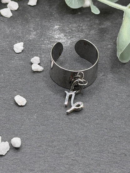 Bague Cassiopée à monture large en acier inoxydable argenté, avec une breloque du signe du Capricorne suspendue.