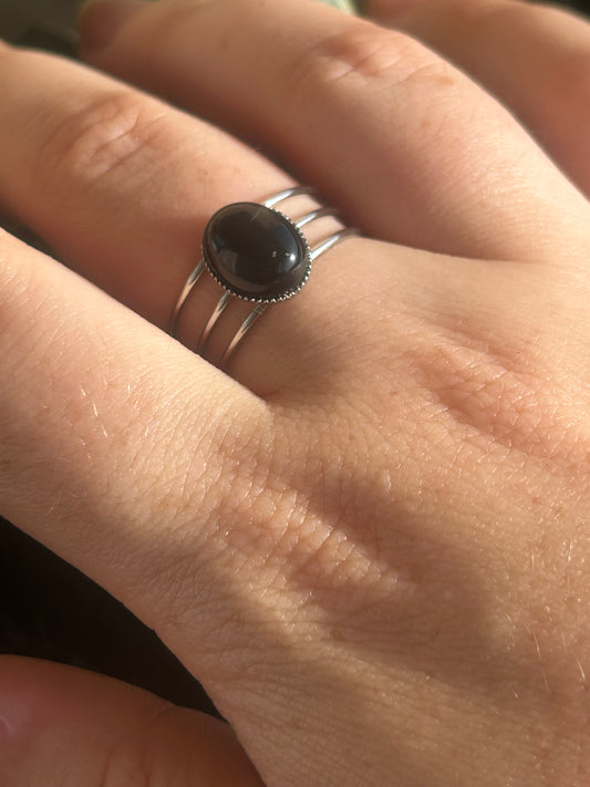 Bague Trinæa à trois anneaux en acier inoxydable argenté et agate noire ovale, portée au doigt.
