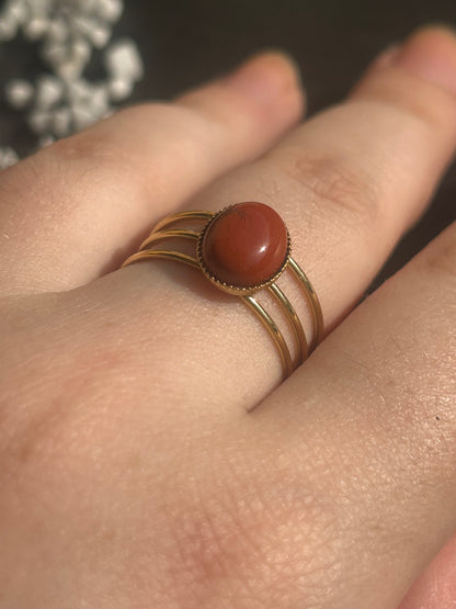 Bague Trinæa à trois anneaux en acier inoxydable doré et jaspe rouge ovale, portée au doigt.