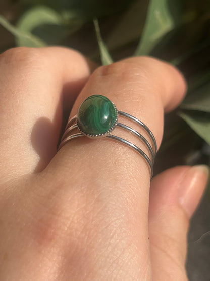 Bague Trinæa à trois anneaux en acier inoxydable argenté et malachite ovale aux motifs verts, portée au doigt.