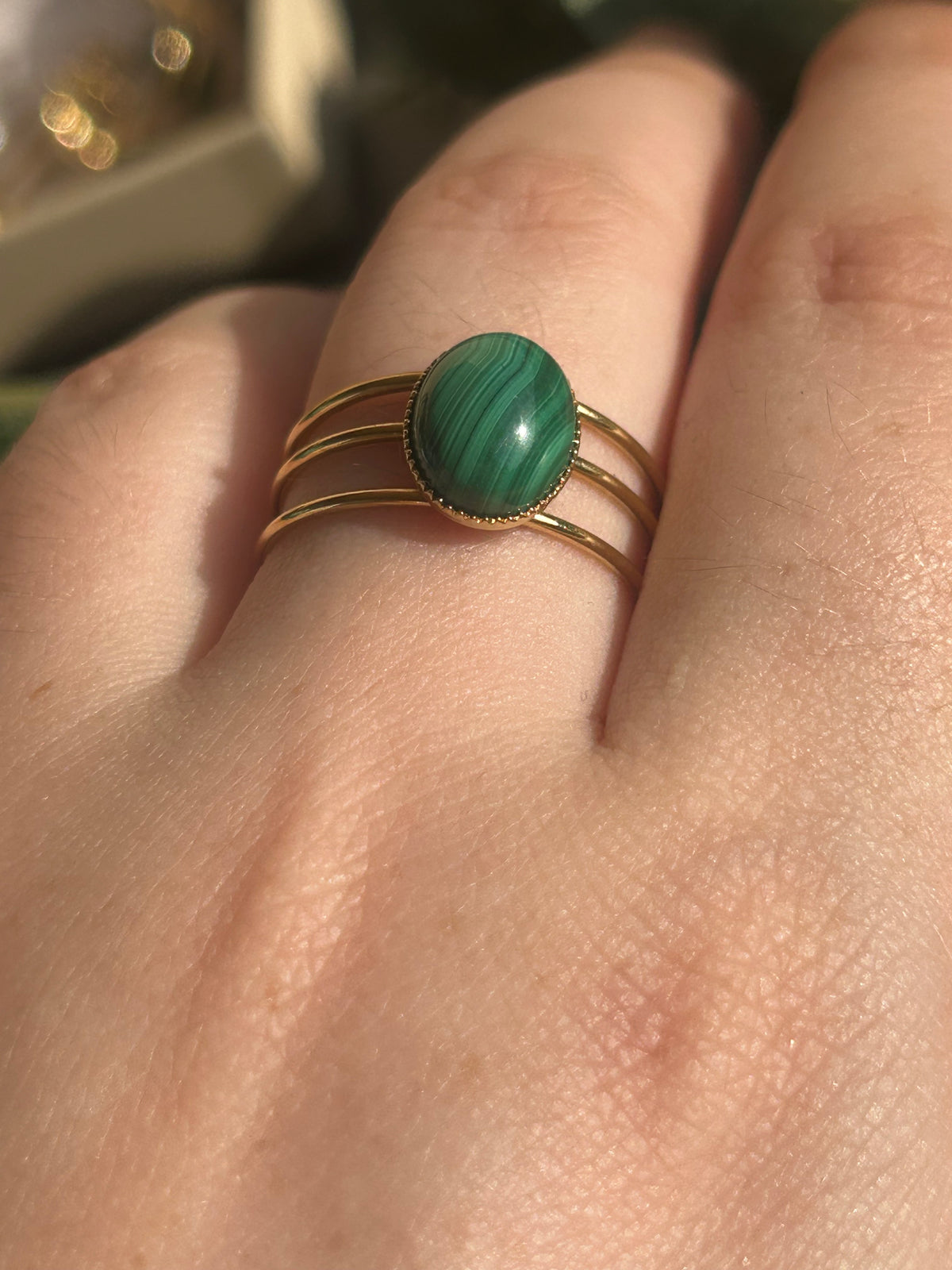 Bague Trinæa à trois anneaux en acier inoxydable doré et malachite ovale aux motifs verts, portée au doigt.