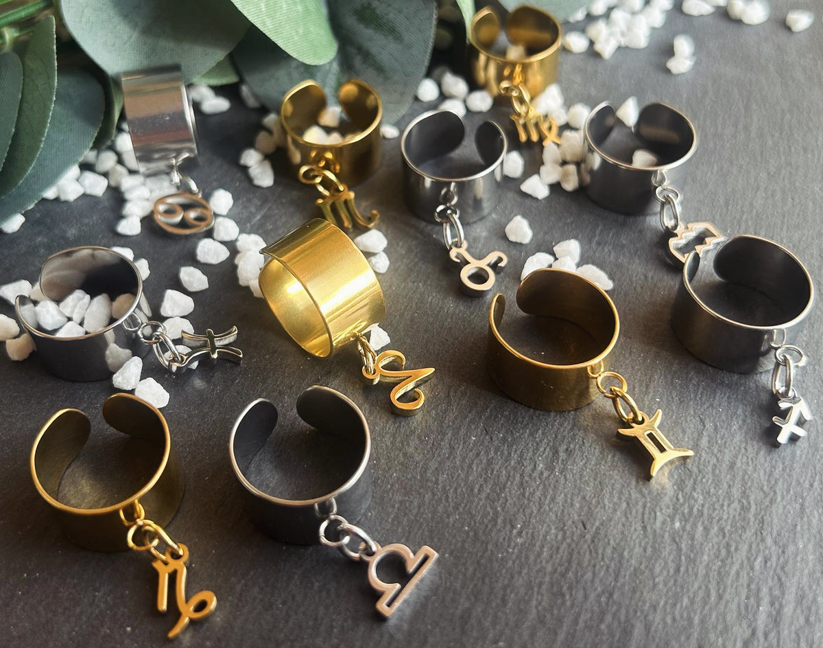 Ensemble de bagues Cassiopée à montures larges en acier inoxydable doré et argenté, avec différentes breloques astrologiques suspendues.
