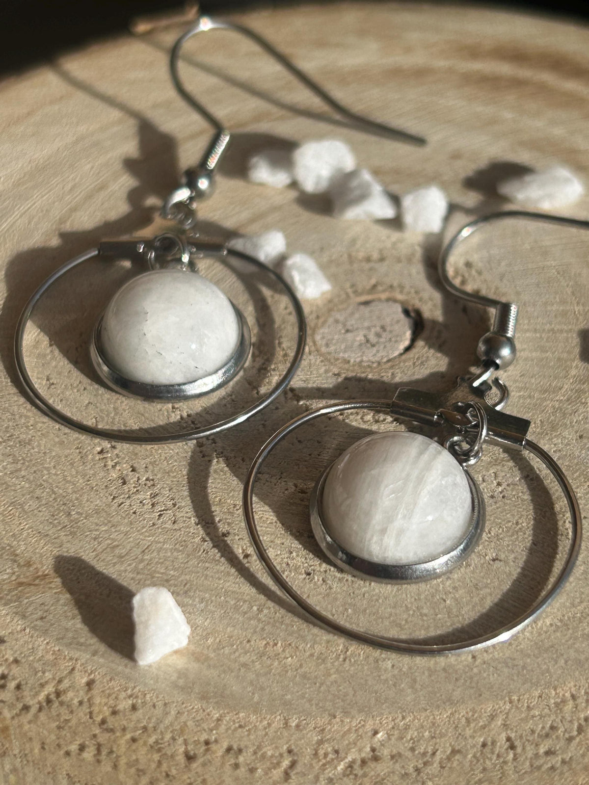 Paire de boucles d’oreilles pendantes Célestia en acier inoxydable argenté, avec des montures circulaires et des cabochons ronds en pierre de lune blanche.